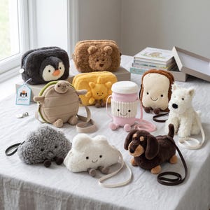 Puede incluir: Una colección de monederos y juguetes de peluche con forma de animales, como un pingüino, un oso, una tortuga, una nube y un perro. Los monederos tienen cremalleras y correas. Los juguetes están colocados sobre una superficie clara.