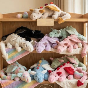 Puede incluir: Una estantería de madera exhibe una colección de juguetes de dragón de peluche en varios colores, incluyendo blanco, negro, rosa y azul. Los dragones tienen alas y texturas suaves. También se ve una manta de arcoíris y huevos decorativos.