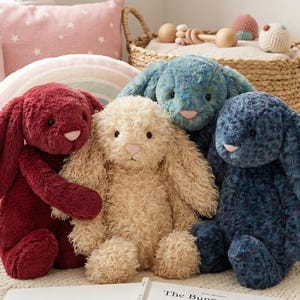 Puede incluir: Cuatro conejos de peluche en tonos rojo, beige, azul y azul marino están dispuestos juntos. Cada conejo tiene largas orejas caídas y una nariz rosada. Una cesta con juguetes y almohadas está en el fondo.