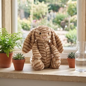 Puede incluir: Un conejito de peluche marrón claro con orejas largas y caídas y una nariz rosada está sentado en un alféizar. El conejito tiene un diseño texturizado y acanalado. Pequeñas plantas en macetas y una jarra de vidrio también están en el alféizar.