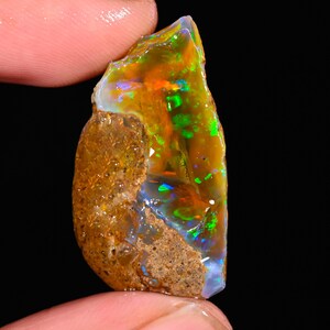 Puede incluir: Una gema de ópalo en bruto con una vibrante exhibición de colores. La piedra presenta una mezcla de tonos naranja, verde y azul, con una superficie rugosa y texturizada. El ópalo se sostiene entre los dedos, mostrando su belleza natural y patrones únicos.