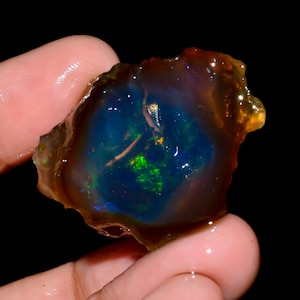 Peut inclure: Une pierre d'opale brute, avec un cœur bleu et vert vif, entouré d'une couche extérieure translucide de couleur ambre. L'opale est tenue dans une main sur un fond noir, mettant en valeur ses couleurs naturelles.