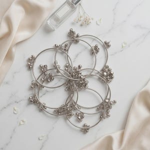 Puede incluir: Una colección de pulseras plateadas con pequeños adornos en forma de campana, dispuestas sobre una superficie de mármol blanco. También hay un frasco de perfume transparente y delicadas flores blancas, junto con una tela de seda color crema.