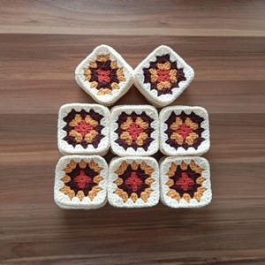 May include: Nine crocheted granny squares arranged on a wooden surface. Each square features a central design in shades of red, orange, and brown, surrounded by a cream-colored border. The squares are handmade and have a textured appearance.