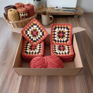 May include: A cardboard box filled with stacks of square, crocheted coasters in shades of red, brown, yellow, and white. A ball of red yarn sits in the box. A basket of crocheted items and craft supplies are in the background.