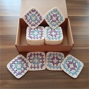May include: A collection of square crochet coasters in a box. Each coaster features a white base with a central floral pattern in light blue and purple. The coasters are stacked and arranged, showcasing their handmade quality.