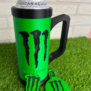 Könnte beinhalten: Ein leuchtend grüner Dosenhalter mit schwarzem Griff und einem schwarzen Monster-Energy-Logo auf der Vorderseite. Der Halter enthält eine silberne Dose, auf der oben der Text AZÚCAR/AÇÚCAR zu sehen ist. Zwei weitere grün-schwarze Logo-Teile liegen daneben.