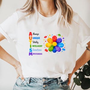 May include: White t-shirt with the word "AUTISM" in colorful letters, each letter representing a positive trait. A flower made of puzzle pieces in rainbow colors is on the shirt.
