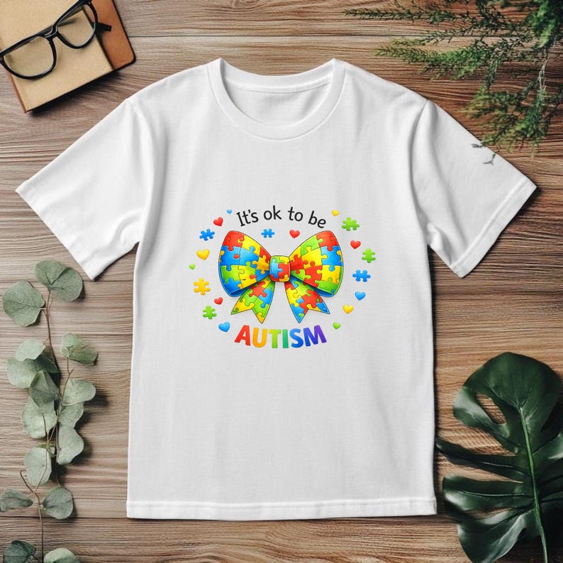 May include: White t-shirt with a colorful bow tie design made of puzzle pieces. The shirt has the text "It's ok to be AUTISM" in a playful font, surrounded by hearts and stars. The shirt is made of a soft material.