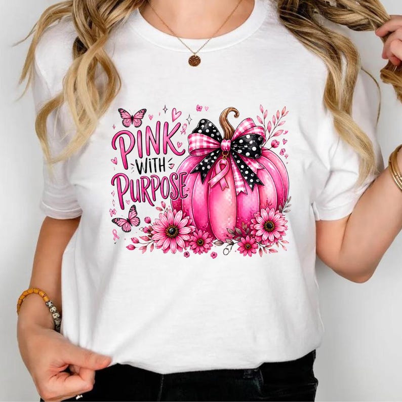 Op de afbeelding: Een wit T-shirt met korte mouwen en een grafische print van een roze pompoen versierd met een zwart-witte strik met stippen en een roze lint. Het ontwerp bevat roze bloemen, vlinders en de tekst "PINK WITH PURPOSE" in het roze.