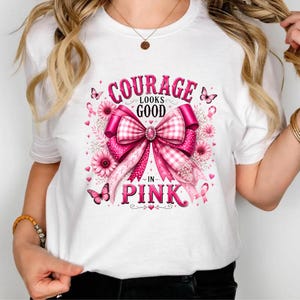 Op de afbeelding: Een wit t-shirt met een grote roze afbeelding van een strik, bloemen en vlinders. De tekst luidt Courage Looks Good In Pink. Het ontwerp bevat een roze lintsymbool, een ruitpatroon op de strik en verschillende tinten roze.
