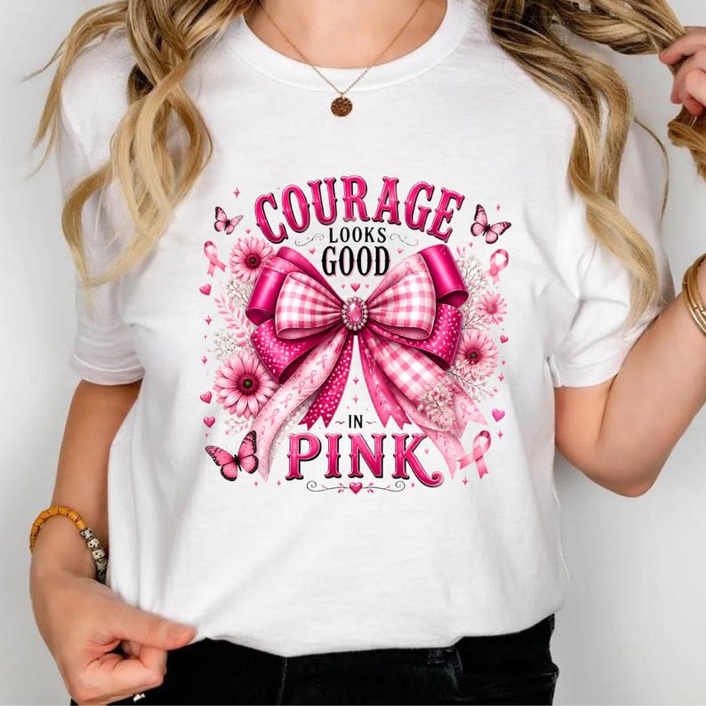 Op de afbeelding: Een wit t-shirt met een grote roze afbeelding van een strik, bloemen en vlinders. De tekst luidt Courage Looks Good In Pink. Het ontwerp bevat een roze lintsymbool, een ruitpatroon op de strik en verschillende tinten roze.