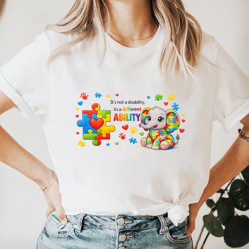 May include: White t-shirt featuring a colorful puzzle piece design with a heart and a cartoon elephant. The text reads "It's not a disability, it's a different ABILITY."