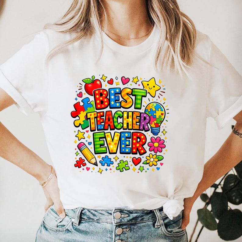 May include: White t-shirt with a colorful graphic that reads "BEST TEACHER EVER." The design includes a red apple, puzzle pieces, stars, a lightbulb, a pencil, and flowers. The shirt is a casual, crew neck style.