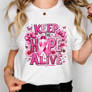 Op de afbeelding: Een wit T-shirt met korte mouwen en een roze grafisch ontwerp met de tekst KEEP THE HOPE ALIVE. Het ontwerp bevat een roze bewustwordingslint, strikken, bloemen en vlinders in verschillende tinten roze met ruitjespatronen en stippen.