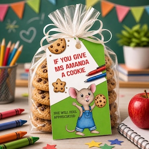 May include: A clear cellophane bag tied with a white ribbon, filled with chocolate chip cookies. A green tag reads "If You Give Ms Amanda A Cookie" with a cartoon mouse holding a cookie. Pens and crayons are visible.