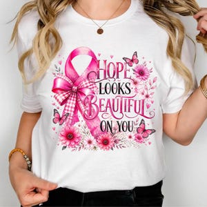 Op de afbeelding: Een wit T-shirt met korte mouwen voorzien van een grote roze bewustwordingslint met een ruitpatroon, omringd door roze bloemen en vlinders. De tekst "HOPE LOOKS BEAUTIFUL ON YOU" staat in een decoratief zwart lettertype in het midden.