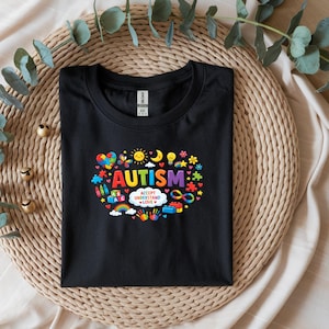 May include: Black crew neck t-shirt with a colorful graphic design. The design includes the word "AUTISM" in rainbow colors, surrounded by various symbols like hearts, puzzle pieces, and a rainbow. The phrase "ACCEPT UNDERSTAND LOVE" is also included.