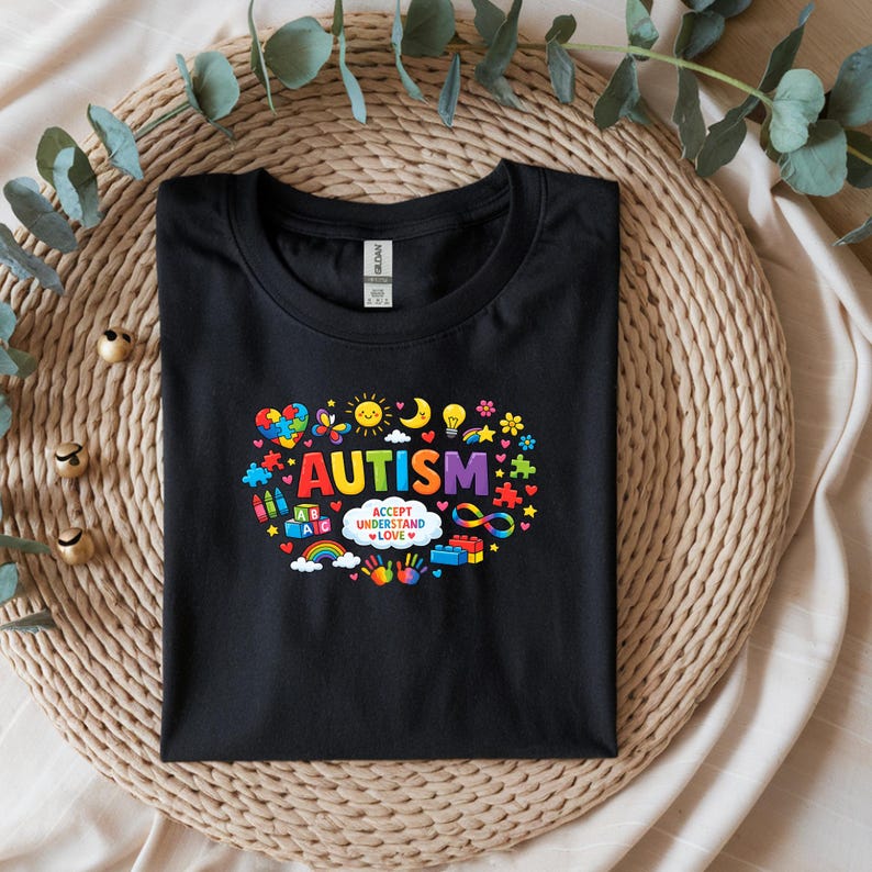 May include: Black crew neck t-shirt with a colorful graphic design. The design includes the word "AUTISM" in rainbow colors, surrounded by various symbols like hearts, puzzle pieces, and a rainbow. The phrase "ACCEPT UNDERSTAND LOVE" is also included.