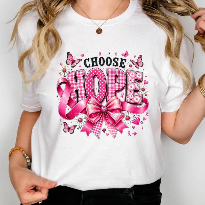 Op de afbeelding: Een wit T-shirt met korte mouwen en een grafische print met de tekst CHOOSE HOPE in zwarte letters. Het ontwerp bevat een grote roze strik, roze vlinders, witte madeliefjes en een roze gingham-patroon op de letters.