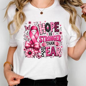 Op de afbeelding: Een wit t-shirt met een grafisch ontwerp met een roze bewustzijnslint, een geruite strik en bloemen. De tekst HOPE IS STRONGER THAN FEAR staat in roze en zwarte letters met ruitpatronen, omringd door kleine vlinders en hartjes in het ontwerp.