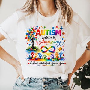 May include: White t-shirt with a colorful graphic design. The design includes the word "AUTISM" above the phrase "Embrace The Amazing", a tree with puzzle piece leaves, and an infinity symbol. The shirt also has the words "Celebrate, Understand, Include, Love".