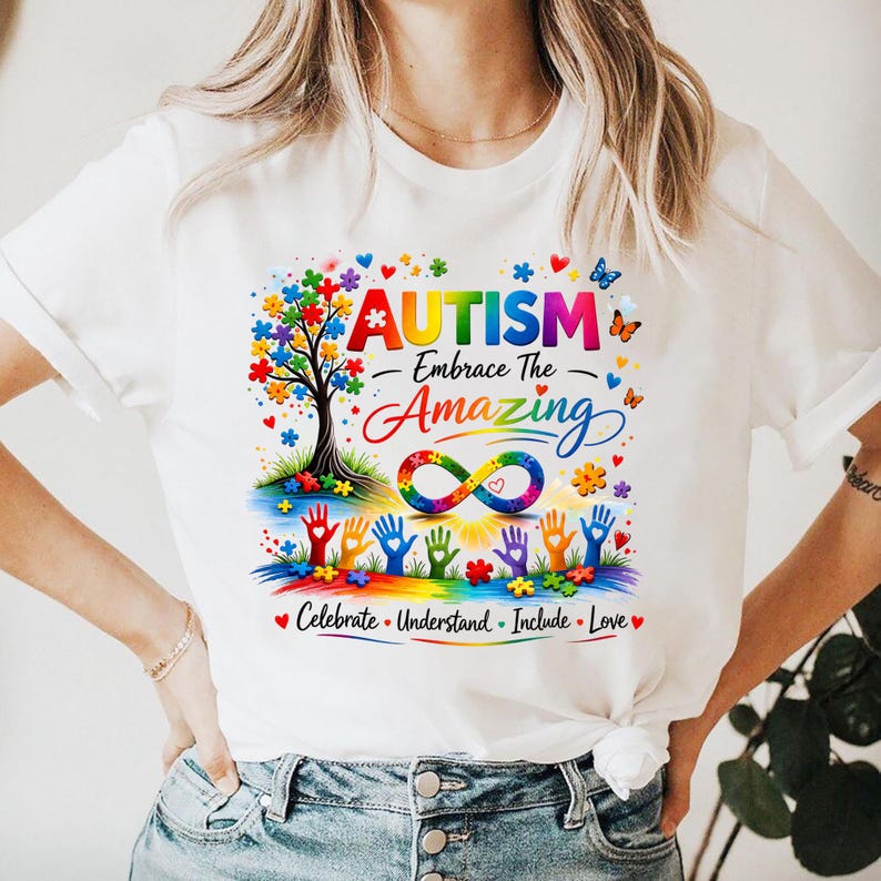 May include: White t-shirt with a colorful graphic design. The design includes the word "AUTISM" above the phrase "Embrace The Amazing", a tree with puzzle piece leaves, and an infinity symbol. The shirt also has the words "Celebrate, Understand, Include, Love".