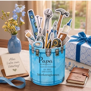 Può includere: Un contenitore blu a forma di cassetta degli attrezzi pieno di ritagli di attrezzi, tra cui un martello, una chiave inglese e una livella. Il contenitore ha la parola "Papa" e testo in tedesco. Sono presenti anche un piccolo vaso di fiori blu e una confezione regalo con un nastro blu.