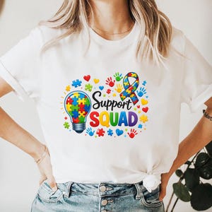 May include: White t-shirt with a colorful graphic design. The design includes a lightbulb made of puzzle pieces, a ribbon, handprints, hearts, and the words "Support Squad" in rainbow colors. The shirt is a casual style.