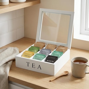 Puede incluir: Una caja de té de madera blanca con tapa de cristal, llena de varias bolsitas de té. La caja lleva la etiqueta "TEA" en letras negras. Las variedades de té incluyen Manzanilla, Té Verde, Earl Grey, Menta y Té Negro. Una taza de té y una cuchara de madera están cerca.