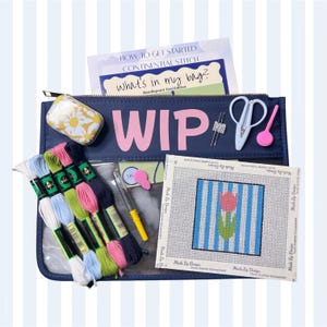 May include: A navy blue zippered pouch with the text WIP in pink letters, containing needlepoint supplies including a white and yellow floral tin, white scissors, a needle threader, a yellow tool, and six skeins of embroidery floss in various colors.