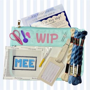 May include: A needlepoint kit with a light blue zippered pouch labeled "WIP" and containing scissors, a needle, and a booklet. The kit includes a canvas with "MEE" stitched in blue, a pen, a luggage tag, and skeins of blue and cream thread.