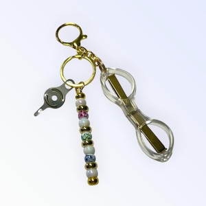 May include: A gold-colored keychain with a clear plastic eyeglasses holder, a small silver tool, and a beaded charm. The charm features white, gold, pink, and green beads. The keychain has a lobster clasp and a key ring.