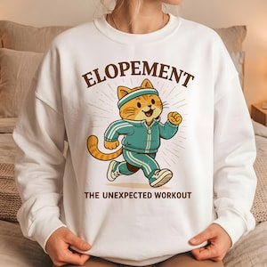 May include: White sweatshirt featuring a cartoon cat in a teal tracksuit, running. The text "ELOPEMENT" is above the cat, and "THE UNEXPECTED WORKOUT" is below. The cat has a headband and is smiling.