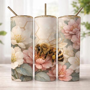 Puede incluir: Un vaso cilíndrico con pajita, que presenta un patrón de flores blancas y rosas, hojas verdes y un diseño de panal dorado. Una ilustración detallada de una abeja está centrada en la superficie, rodeada de pétalos de flores suaves.