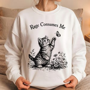 May include: White sweatshirt with a black and white graphic of a kitten reaching for a butterfly. The text "Rage Consumes Me" is arched above the kitten. The design includes flowers and grass.