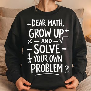 May include: Black sweatshirt with the text "+ DEAR MATH, GROW UP, X - AND - √, - SOLVE = 1/3 YOUR OWN PROBLEM?" in white. The sweatshirt has a crew neck and long sleeves.