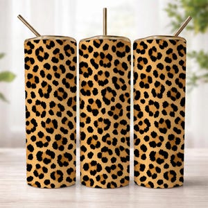 May include: Three stainless steel tumblers with a leopard print design. Each tumbler has a gold-colored lid and a matching straw. The tumblers are cylindrical and stand upright on a light-colored surface.