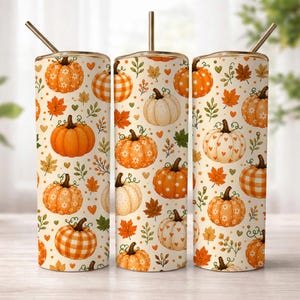 Op de afbeelding: Drie cilindrische bekers met deksels en rietjes, voorzien van een herhalend patroon van oranje en witte pompoenen, herfstbladeren en kleine hartjes op een crèmekleurige achtergrond. De pompoenen tonen diverse texturen zoals ruitjes, stippen en bloemmotieven.