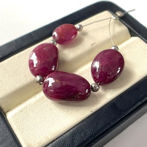 May include: A strand of four polished, deep red oval-shaped ruby gemstones separated by small, round, metallic silver-coloured spacer beads. The stones are strung on a thin, flexible wire and arranged in a curved formation on a display base.