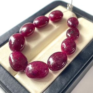 Peut inclure: Un collier de dix pierres précieuses rubis polies de forme ovale, d'une teinte rouge magenta profond. Les pierres sont enfilées sur un fil transparent fin et disposées en courbe, révélant des inclusions naturelles et une texture lisse et brillante.