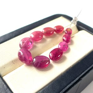 Op de afbeelding: Een streng van tien gepolijste, ovaalvormige fuchsia roze edelstenen gerangschikt in een druppelvormige lus. De kralen variëren enigszins in grootte en zijn geregen aan een dun, doorzichtig draadje, met een levendige kleur en gladde oppervlakken.