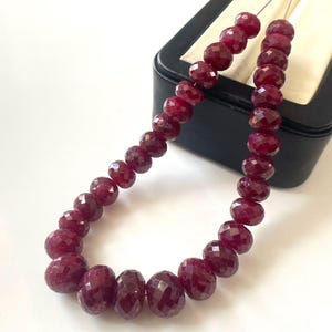 Puede incluir: Un hilo de cuentas de piedra preciosa de color rojo intenso, facetadas y redondas, dispuestas en una línea curva. Las cuentas exhiben una calidad translúcida con una superficie pulida, mostrando un color y textura consistentes en toda la longitud.