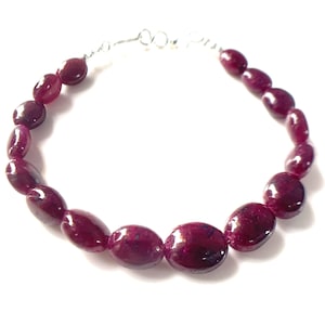 Op de afbeelding: Een armband bestaande uit gepolijste, ovaalvormige diepmagenta kralen. De kralen zijn in een cirkelvormige opstelling geregen en aan de bovenkant verbonden door een dunne, zilverkleurige metalen draadsluiting, wat een doorlopende lus van gladde stenen vormt.