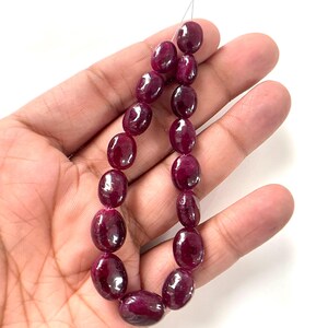 May include: A strand of smooth, polished, oval-shaped ruby beads in a deep, saturated magenta-red color. The beads are strung on a thin, light-colored thread, showcasing a glossy, reflective surface with natural variations in the stone's deep red hue.