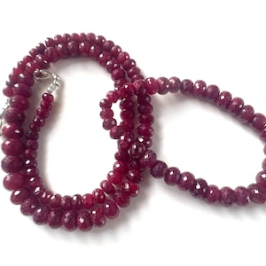 May include: A necklace composed of numerous deep red, faceted, circular beads strung together. The strand features a metallic silver-toned clasp mechanism at one end, allowing for secure fastening. The beads exhibit a consistent dark crimson hue.