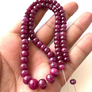 Puede incluir: Un hilo de cuentas de piedras preciosas de color magenta intenso, redondas, lisas y pulidas. Las cuentas están ensartadas en un alambre fino y flexible, mostrando una forma circular consistente y un color rico y saturado en toda la longitud.