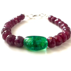 May include: A beaded bracelet featuring a series of faceted, deep red ruby beads arranged in a circular pattern, centered with a single, smooth, oval-shaped vibrant green emerald bead. The bracelet is secured with a silver-toned metal clasp.
