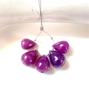 May include: A strand of five polished, teardrop-shaped magenta gemstone beads. The beads are strung on a thin, metallic wire and are separated by small, round silver-colored spacer beads. The stones exhibit a smooth, glossy surface texture.