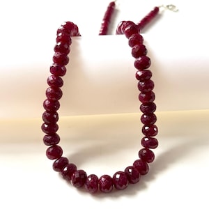 Peut inclure: Un collier composé de perles circulaires facettées d'un rouge profond disposées en un seul rang. Les perles présentent une surface polie et réfléchissante avec une teinte cramoisie sombre constante. Un fermoir en métal argenté est visible.
